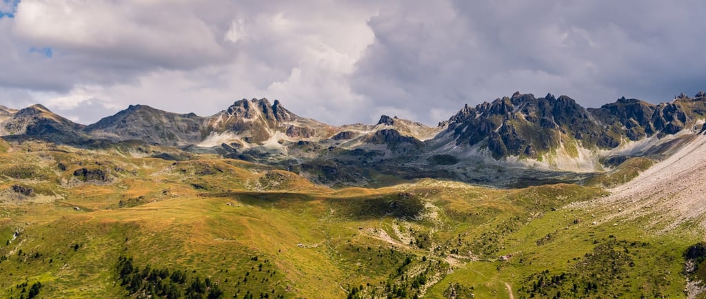 Panoramic view of jagged alpine mountain peaks and green valley slopes under a cloudy sky.