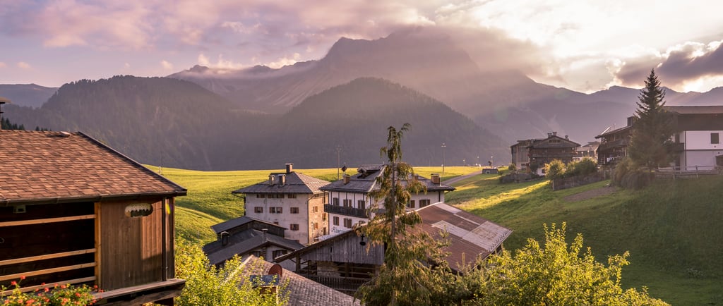 Sunset over a rustic alpine village in the Dolomite Mountains with wooden chalets and green hills.