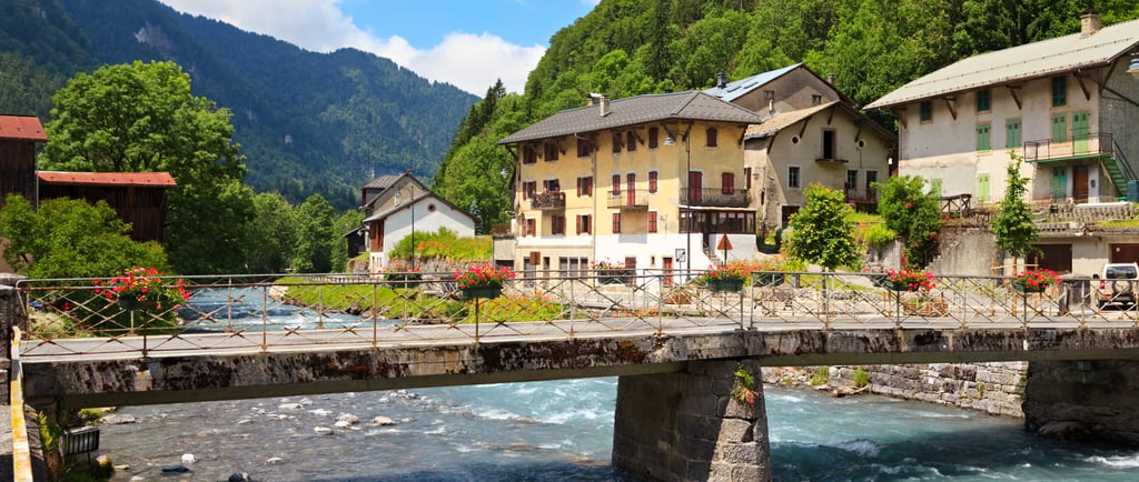 A stone bridge with flowers spans a turquoise river in a scenic French mountain village.