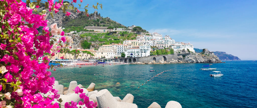 Panoramic view of Amalfi coast with pink bougainvillea flowers, white buildings, and blue sea.