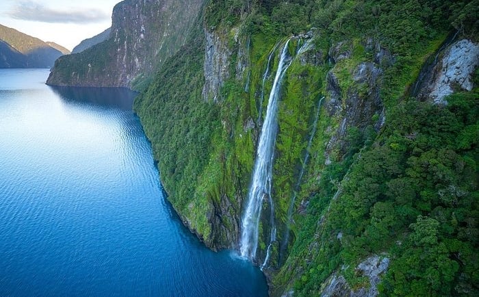 Aerial view of a tall waterfall cascading down lush green cliffs into the blue waters of Milford Sound, New Zealand.