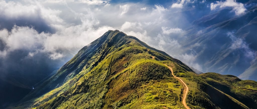 Aerial view of a winding dirt path on a green mountain ridge surrounded by clouds and sunlit peaks.