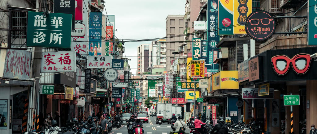 Busy city street in Taiwan with colorful business signs, parked scooters, and commuters on motorcycles.