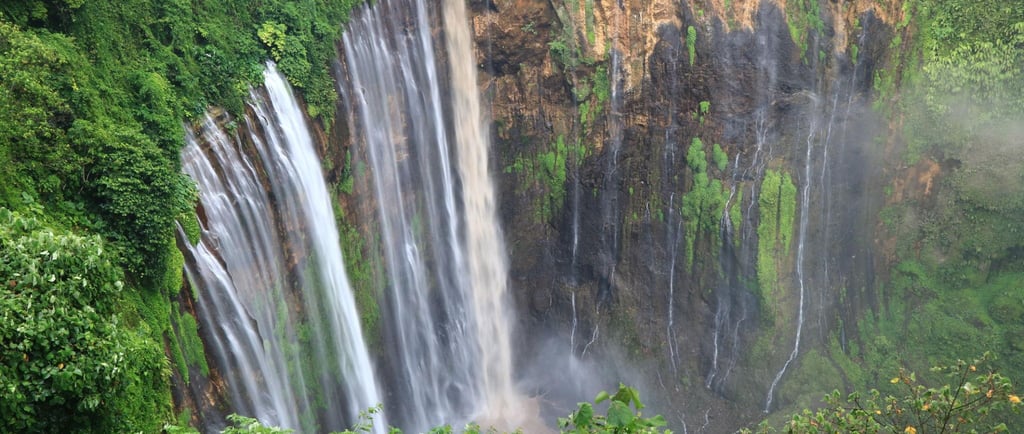 Panoramic view of Tumpak Sewu Waterfall in Indonesia surrounded by lush green tropical jungle.