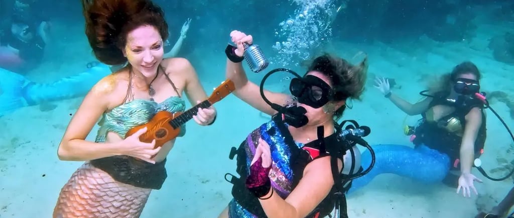 Mermaid and scuba divers performing a concert underwater with a ukulele and microphone on the ocean floor.