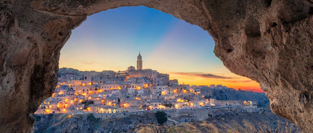 Illuminated ancient city of Matera, Italy, at sunset viewed through a stone cave opening.