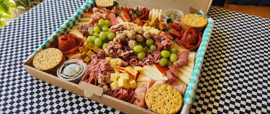 Gourmet charcuterie box with assortment of cured meats, cheeses, grapes, and crackers on a checkered tablecloth.