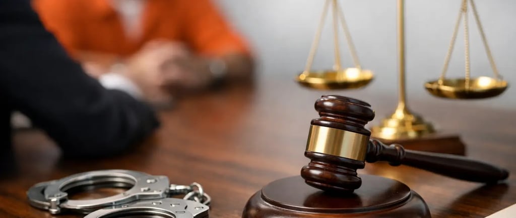 Judge's gavel and handcuffs on a courtroom desk with a defendant in an orange jumpsuit.