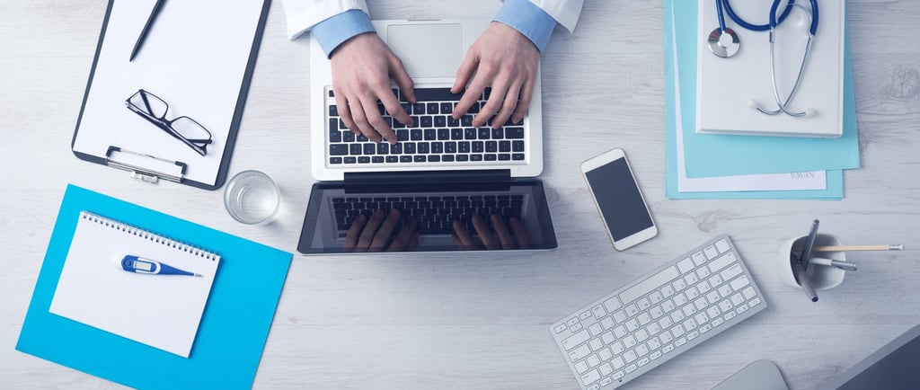 médico-usando-o-computador-em-uma-mesa-onde-estão-celular-papéis-e-objetos-de-trabalho