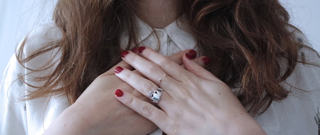 A white woman with brown hair clasping her hands over her heart. She's wearing a white blouse and her nails are painted red.