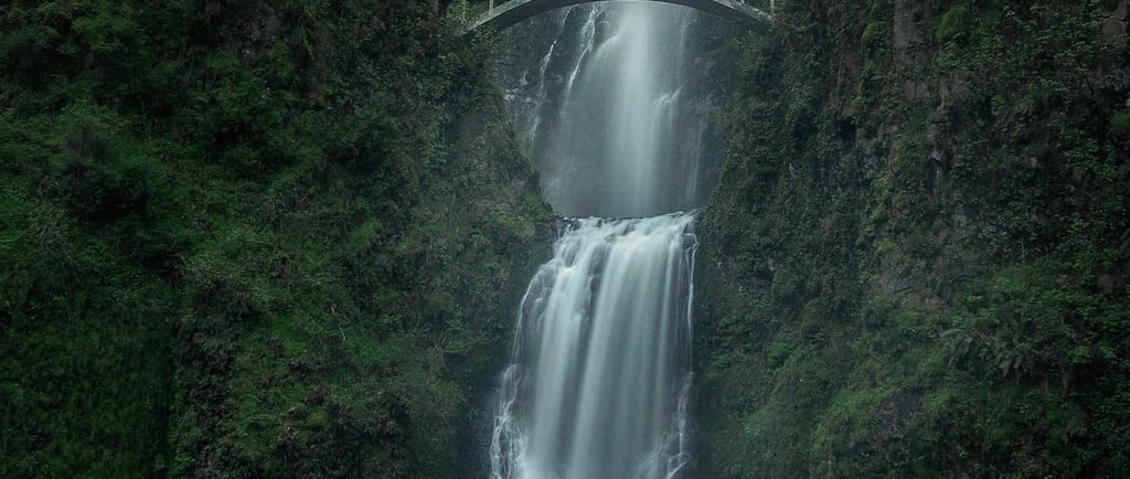 a waterfall cascading down a green gorge and under a walking bridge