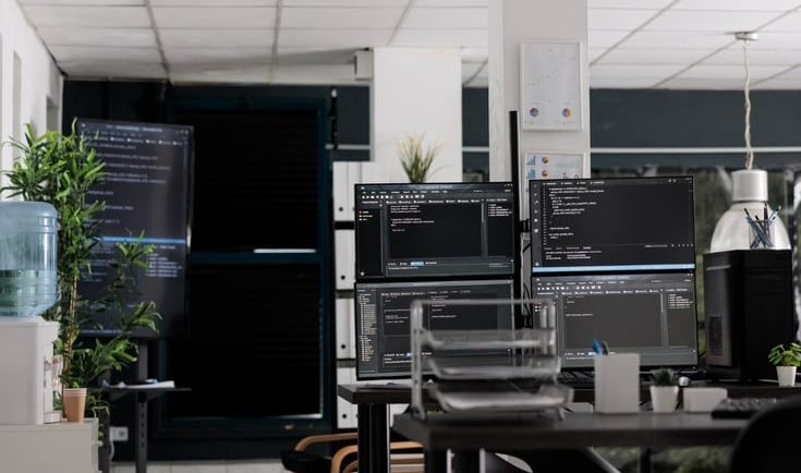 Kantor pengembangan perangkat lunak modern dengan beberapa monitor yang menampilkan kode, melambangkan lingkungan kerja softw