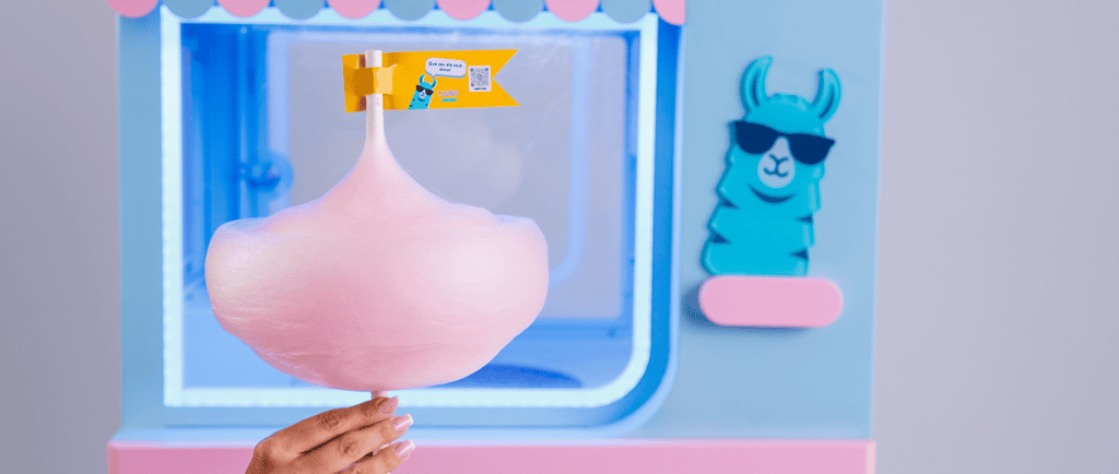 Mão segurando um algodão-doce rosa em frente a uma máquina automática de doces azul