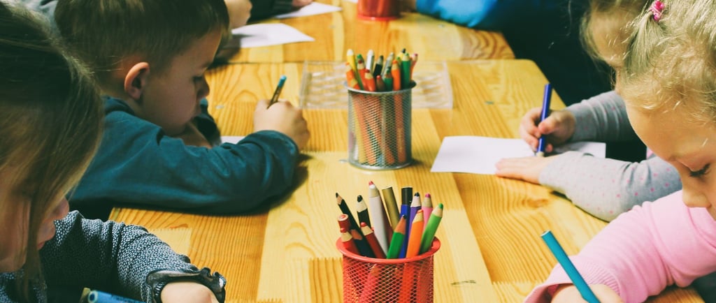 Children are gathered around a wooden table, drawing with colored pencils. 