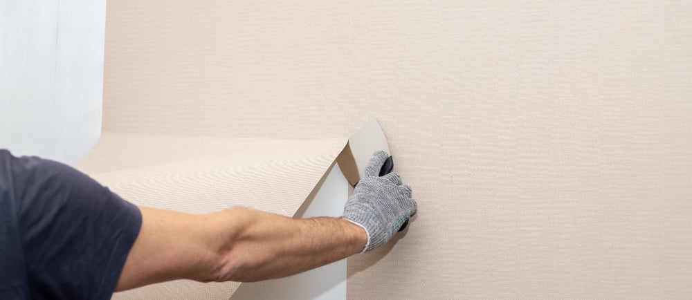 A worker's gloved hand carefully peeling or hanging textured beige wallpaper on a wall during home renovation.