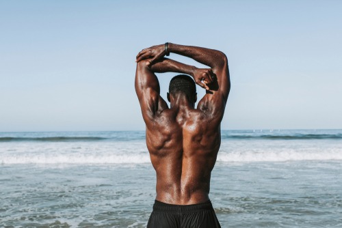 Muscular man stretching his triceps on a sunny beach after an outdoor workout session.