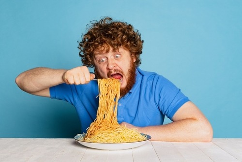 A hungry man with curly red hair eating a large plate of spaghetti noodles against a blue background.