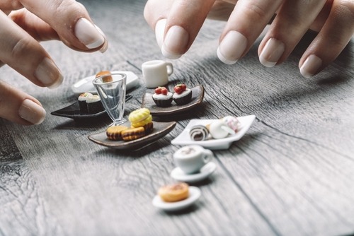 A person with manicured fingernails arranging realistic miniature food items and tiny dishes on a wooden surface.