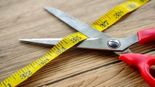 Red handled sewing scissors cutting a yellow measuring tape on a wooden table.
