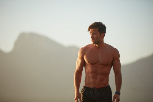 Shirtless muscular man with defined abs training outdoors in front of mountains.
