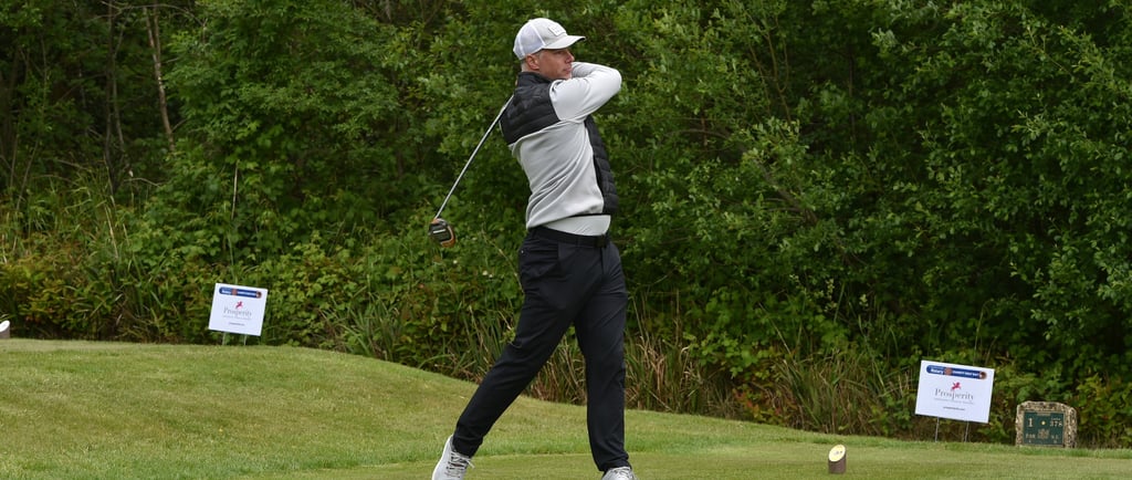 A male golfer in a white cap follows through on a drive during a tournament at a lush green golf course.