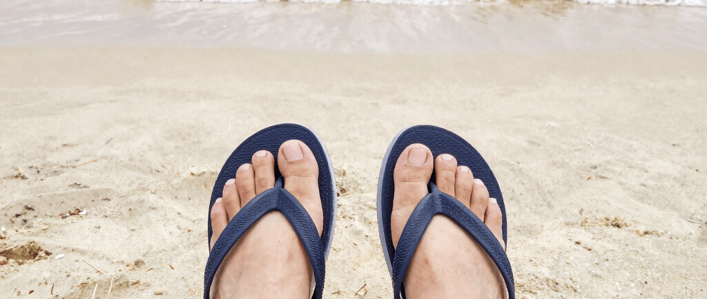 Person wearing navy blue flip flops on a sandy tropical beach with ocean waves.