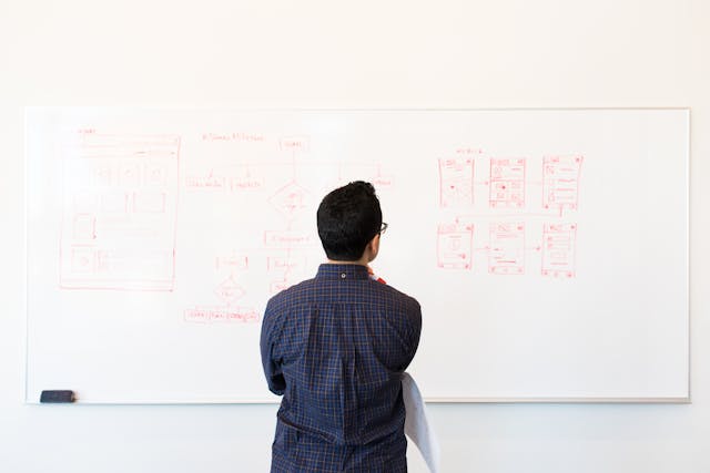 A man looking at a whiteboard displaying a wireframes drawn in red ink.