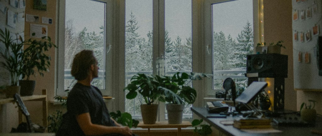 Man sitting in a softly lit room, looking out snowy windows in quiet reflection.