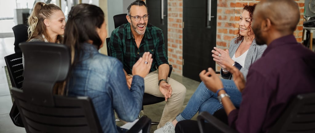 A diverse group of adults sit in a circle, smiling and talking during a relaxed group discussion or