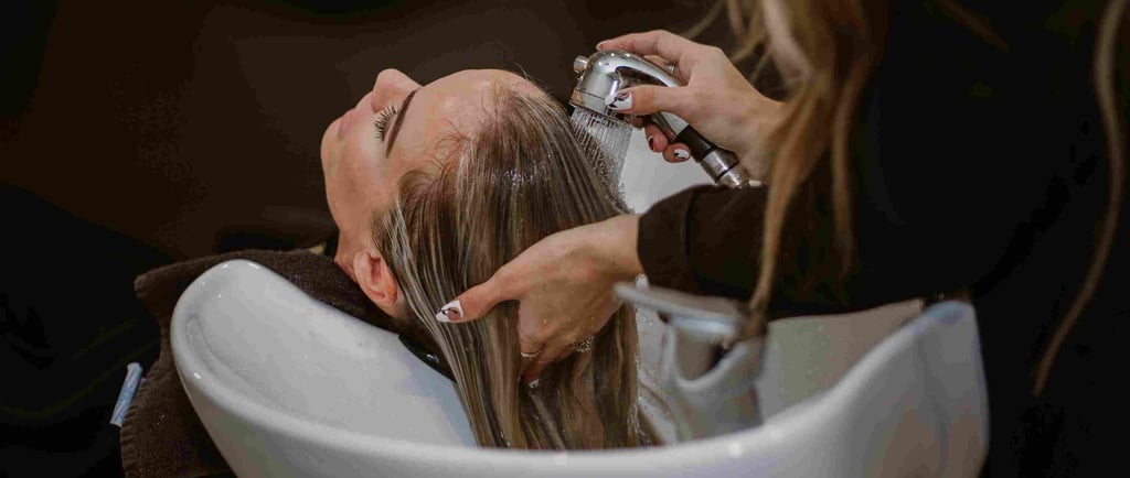 A hair stylist washes a blonde woman's hair in a white salon sink basin using a professional sprayer.