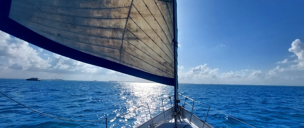 A sailboat sailing on bright blue ocean water under a sunny sky with sun glinting off waves.