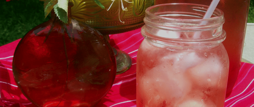 Mason jar with a fruity iced tea and a straw with green and red glass vases on a red tablecloth.