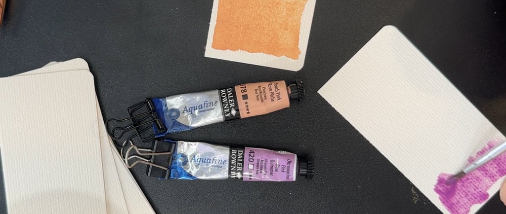 Watercolor paint tubes, watercolor paper, and someone painting on a piece of paper. black background