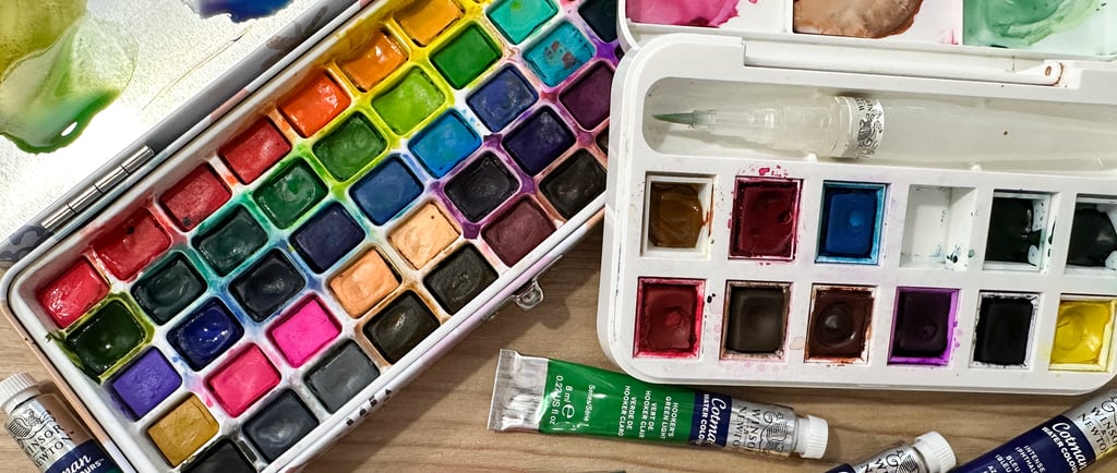Overhead view of watercolor paint palettes, paint tubes, and brushes arranged on a wooden table — essential supplies for lear