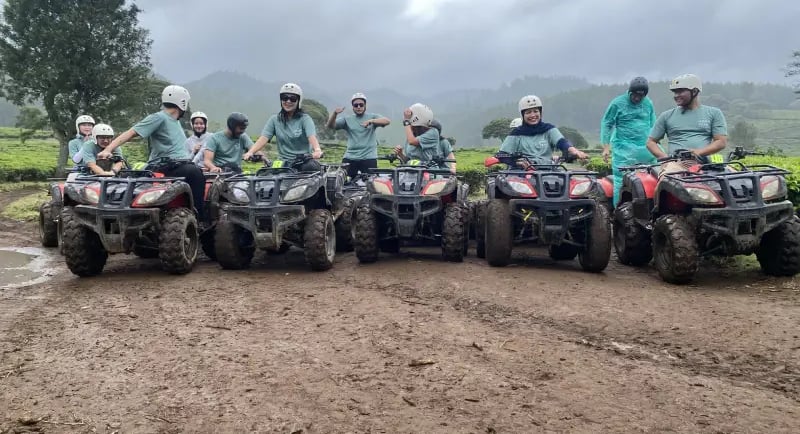 Sekelompok orang dewasa mengendarai ATV di jalur tanah berlumpur di tengah perkebunan teh yang luas.