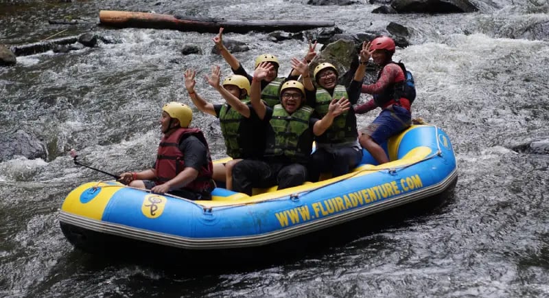 Sekelompok orang ceria sedang melakukan arung jeram di sungai berjeram, mengenakan helm kuning dan jaket pelampung, sambil me