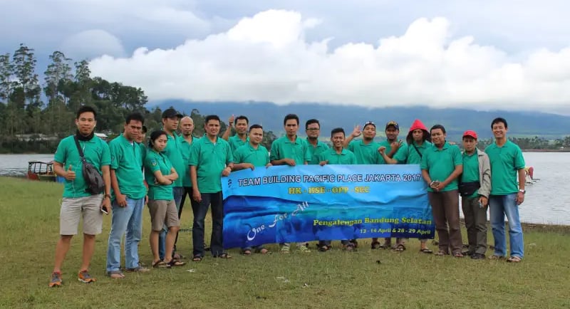 Sekelompok karyawan yang mengenakan kaus hijau kompak berpose di samping spanduk bertuliskan "TEAM BUILDING PACIFIC PLACE JAK