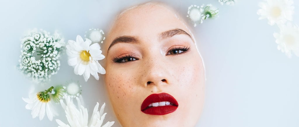 In einer Badewanne mit Milchwasser schaut ein Frauengesicht heraus, um sie herum schwimmen Blumen