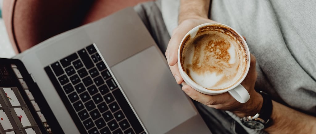an image of man with a laptop on his lap sitting on a couch holding a cup of coffee in a coffee shop