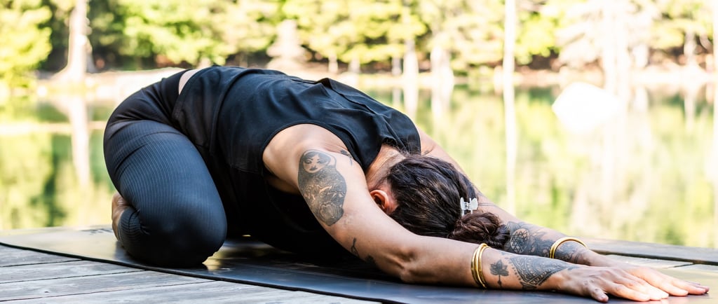 Tattooed woman practicing child's pose yoga on a wooden dock by a calm lake in the forest.