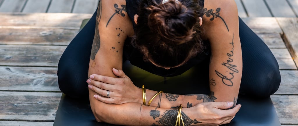 A woman with arm tattoos practicing child's pose on a yoga mat outdoors on a wooden deck.