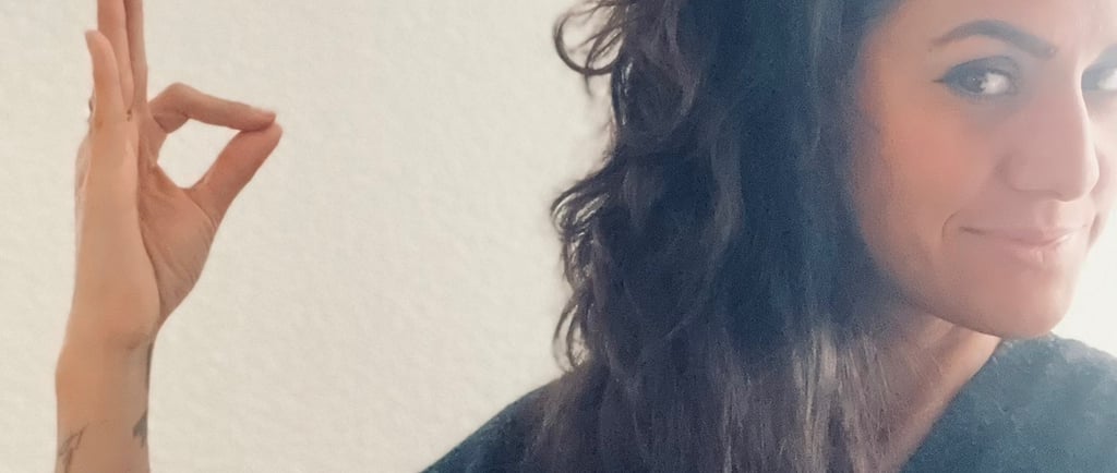 A woman with wavy dark hair and tattoos making an okay hand gesture against a white wall.