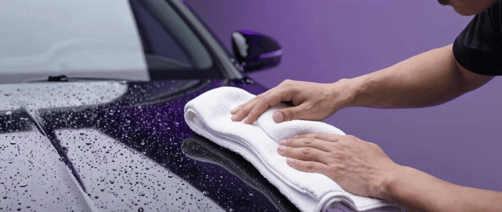 A detailer uses a white microfiber towel to dry a black Mercedes-Benz after a professional car wash.