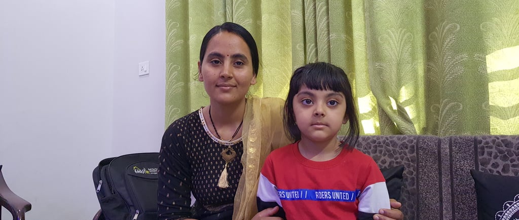 A mother and young daughter sit together on a couch in front of green curtains.