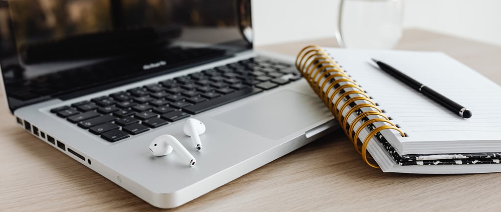 Open laptop with air pods on a desk with notebooks, pen, and glass of water. 