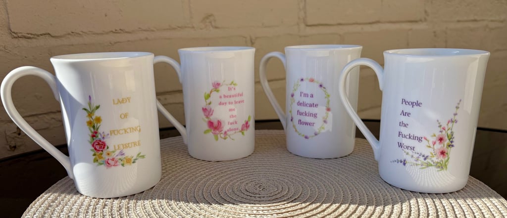 Four white bone china mugs featuring floral designs and humorous, irreverent quotes.