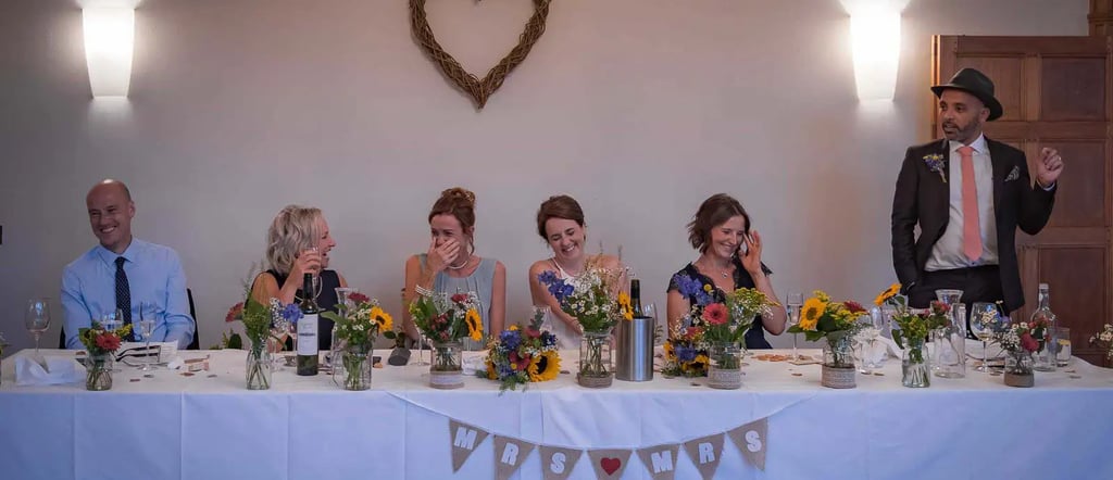 Wedding guests laugh during speeches at a rustic head table decorated with sunflowers and a wicker heart.