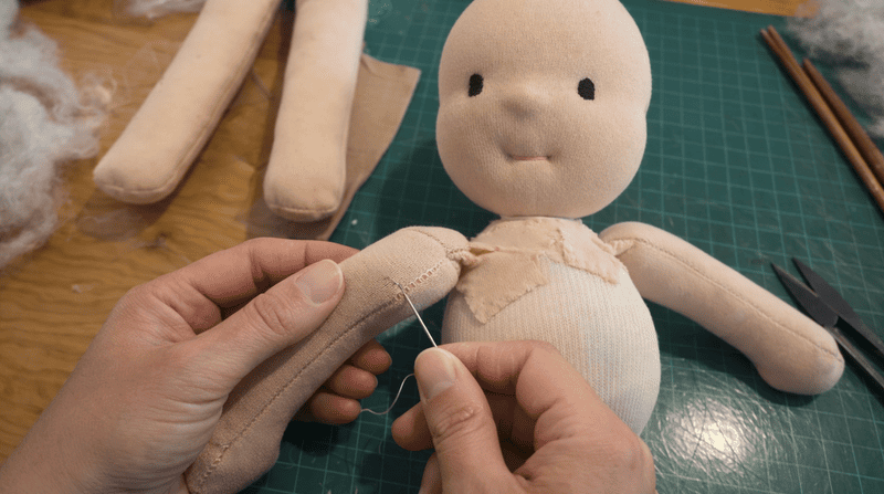 Manos cosiendo brazos de una muñeca y al fondo se ve la cabeza y parte del pecho de una muñeca waldorf en proceso