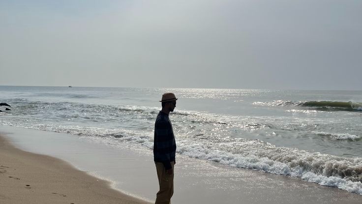Man in a straw hat walking along a sunny beach with ocean waves crashing on the shore.