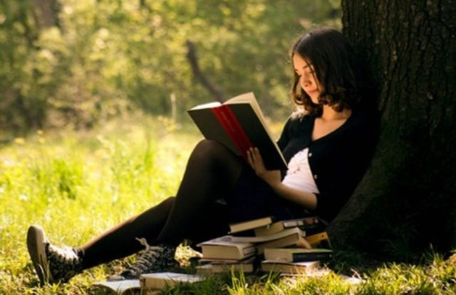 A woman sitting on the ground at the foot of a tree reading a book
